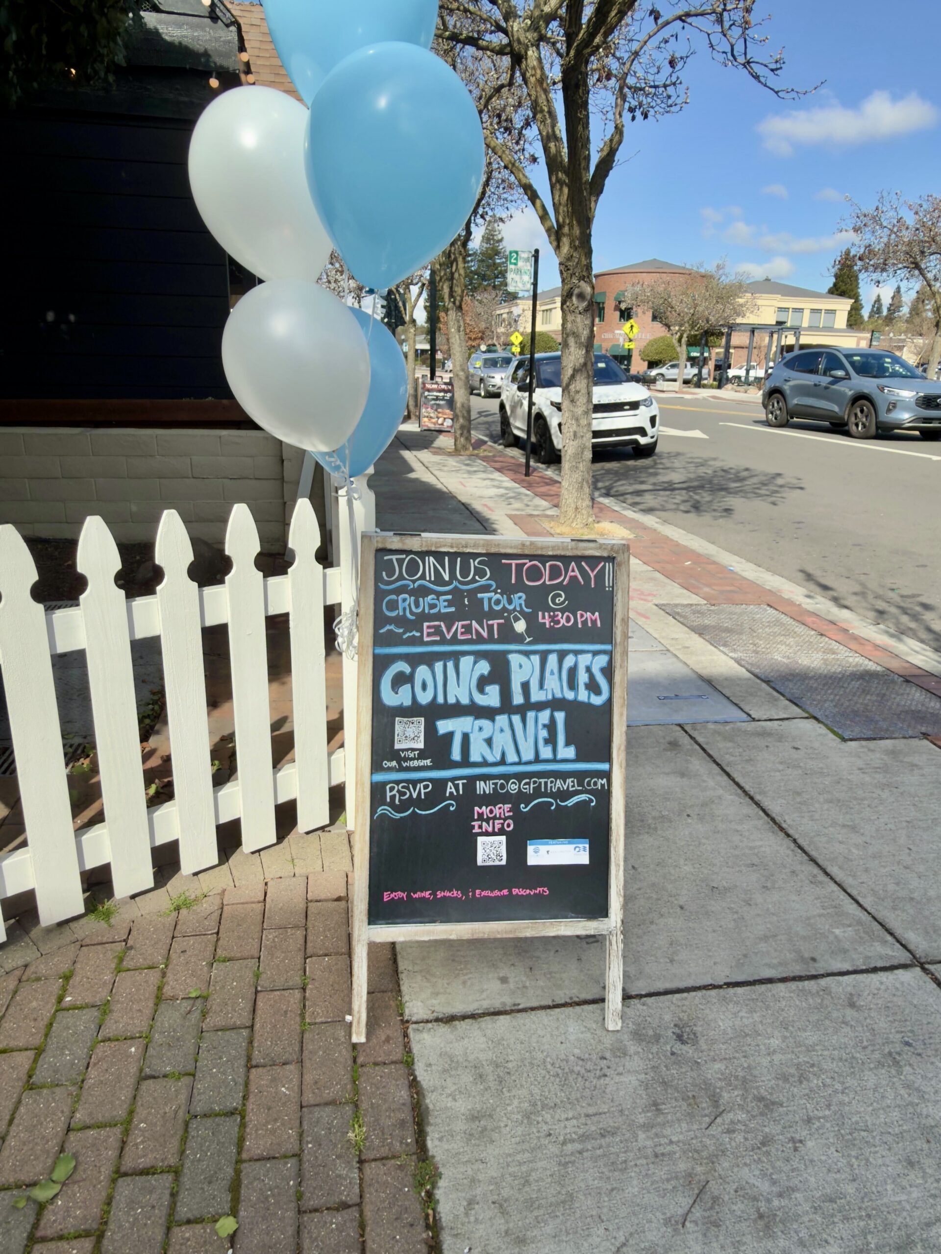 A chalkboard sandwich board sign on a sunny sidewalk decorated with blue and white balloons, advertising a "Going Places Travel" travel event.