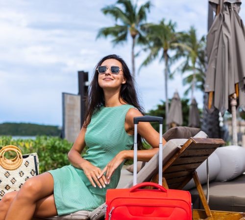 happy-caucasian-pretty-long-hair-elegant-tourist-woman-dress-with-red-suitcase-outside-hotel